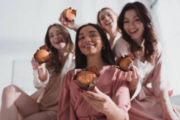 Concentration sélective des femmes multiculturelles montrant des muffins, souriant et regardant la caméra à la fête de célibataire — Photo de stock