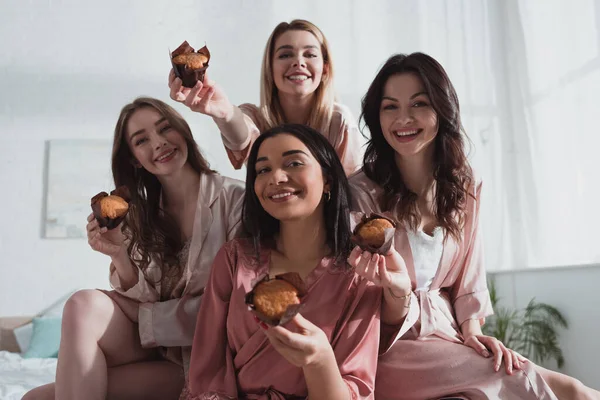 Concentration sélective des femmes multiculturelles souriant et montrant des muffins dans la chambre à la fête de célibataire — Photo de stock