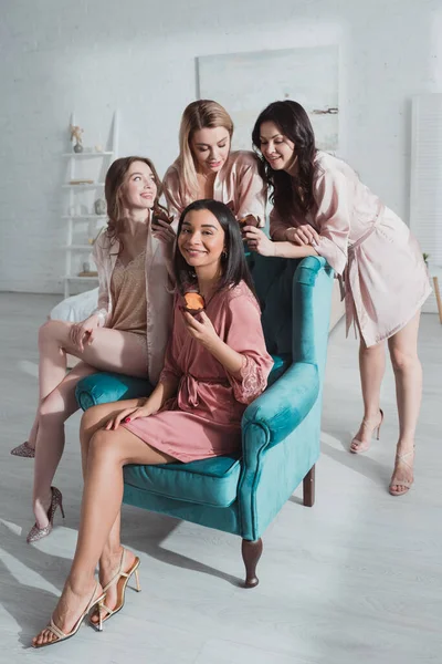 Enfoque selectivo de mujeres multiétnicas con magdalenas felices juntas en sillón en la habitación en la despedida de soltera - foto de stock