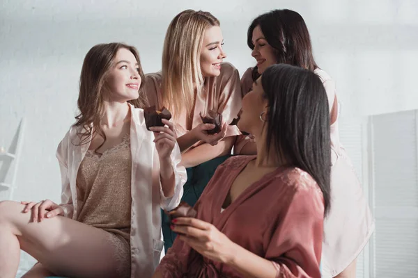Vista de ángulo bajo de las mujeres multiculturales con magdalenas mirándose y sonriendo en la habitación en la despedida de soltera - foto de stock