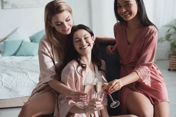 Foyer sélectif de femmes multiethniques cliquetis avec des verres de champagne dans le fauteuil dans la chambre à enterrement de vie de jeune fille — Photo de stock