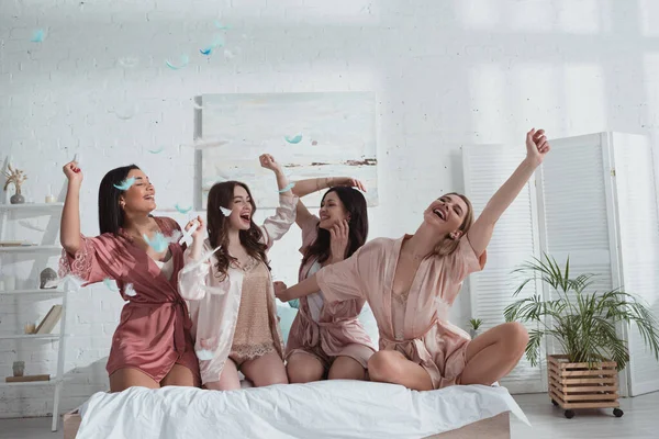 Femmes multiculturelles heureuses et excitées avec les mains dans l'air sur le lit avec des plumes dans la chambre au enterrement de vie de jeune fille — Photo de stock