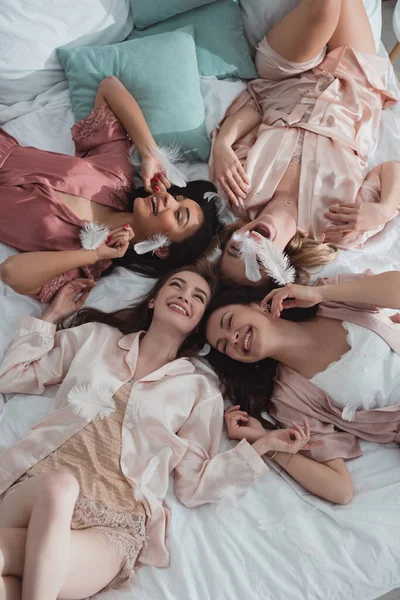 Top view of happy and excited multiethnic women resting on bed with feathers at bachelorette party — Stock Photo