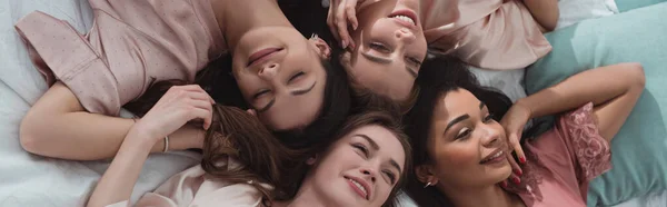 Top view of multiethnic women smiling and resting on bed at bachelorette party, panoramic shot — Stock Photo