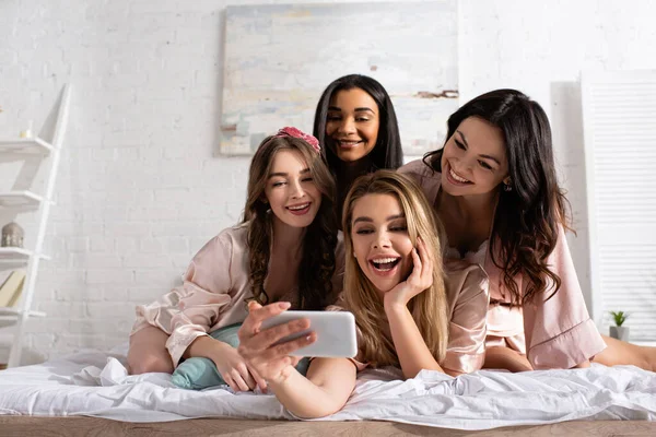 Amigos multiculturales sonriendo y tomando selfie juntos en la cama en la despedida de soltera - foto de stock