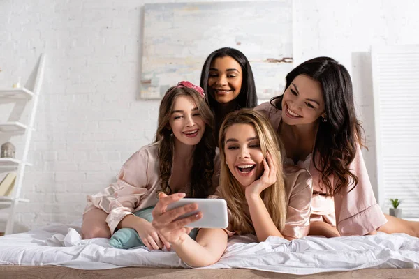 Amis multiculturels souriant et prenant selfie ensemble sur le lit au enterrement de vie de jeune fille — Photo de stock