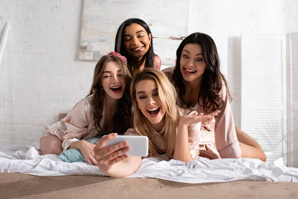 Happy and excited multiethnic women taking selfie on bed at bachelorette party — Stock Photo