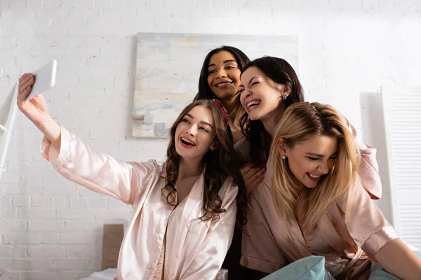 Happy multiethnic friends smiling and taking selfie on bed at bachelorette party — Stock Photo