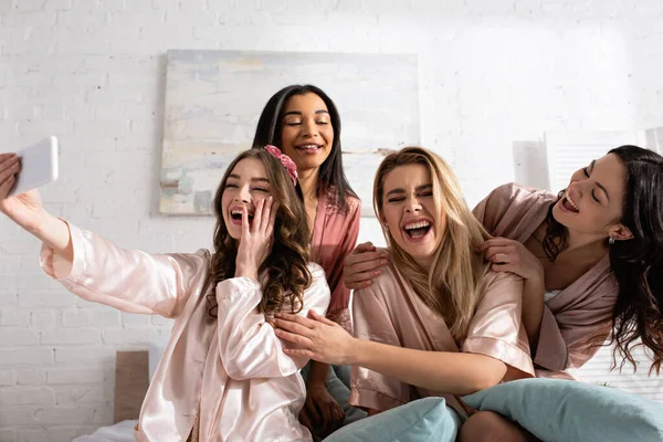 Amigos multiétnicos felizes e animados sorrindo e tomando selfie juntos na cama na festa de despedida de solteira — Fotografia de Stock