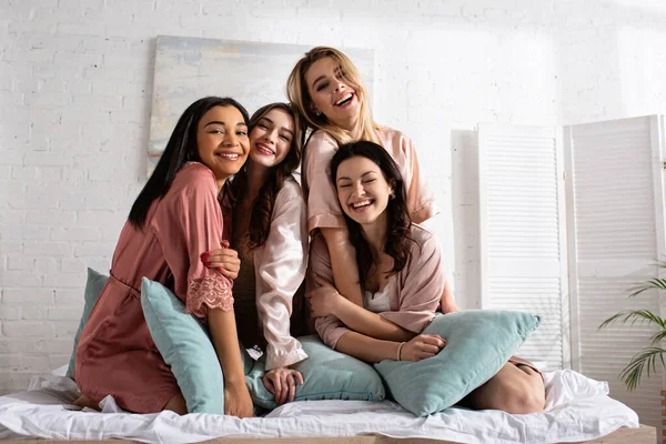 Happy multiethnic friends looking at camera and smiling together with pillows on bed at bachelorette party — Stock Photo