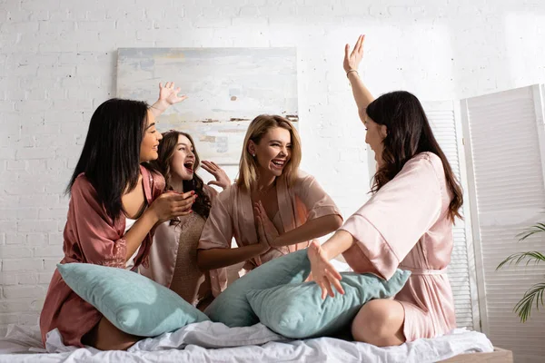 Ami multiethnique excité se regardant souriant et heureux avec des oreillers sur le lit au enterrement de vie de jeune fille — Photo de stock