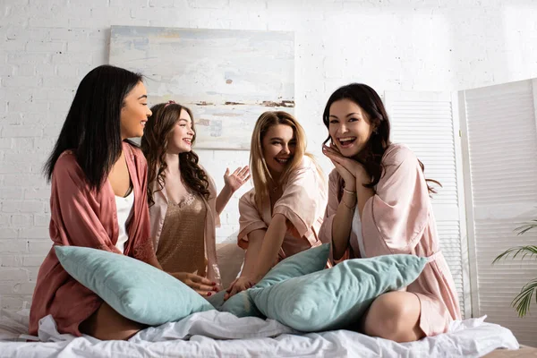 Mulheres multiculturais excitadas sorrindo junto com almofadas na cama na despedida de solteira — Fotografia de Stock