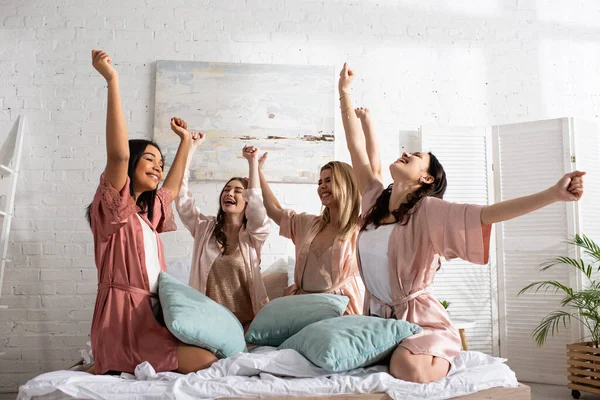 Felices amigos multiculturales con las manos en el aire sonriendo con almohadas en la cama en la despedida de soltera - foto de stock