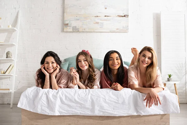 Femmes multiethniques souriantes, regardant la caméra et se reposant sur le lit à la fête de célibataire — Photo de stock