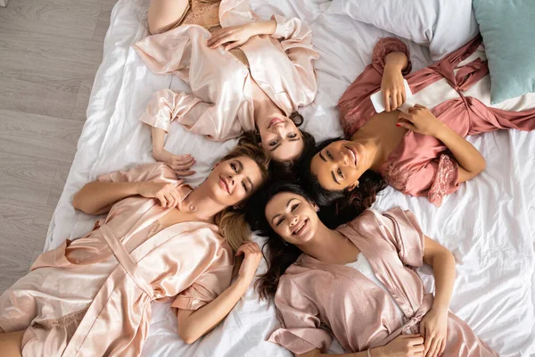 Vue du dessus des femmes multiethniques souriant, regardant la caméra et couché sur le lit à la fête de célibataire — Photo de stock