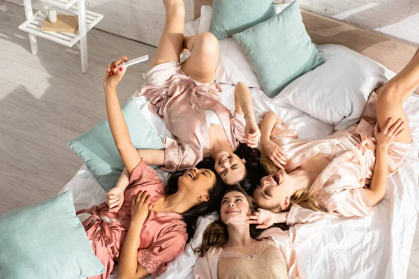 Top view of multiethnic women smiling, taking selfie and lying on bed at bachelorette party — Stock Photo