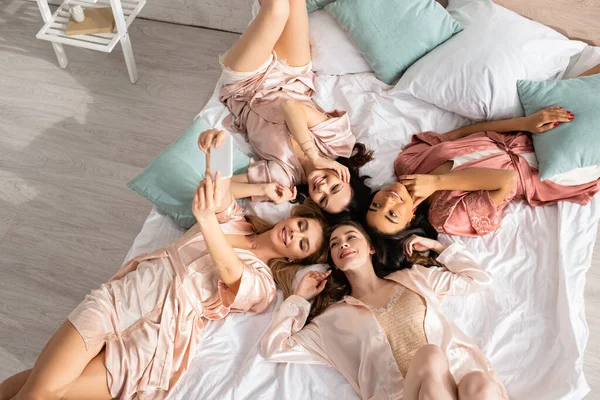 Vista dall'alto di donne multietniche che sorridono, si fanno selfie e si riposano sul letto all'addio al nubilato — Foto stock