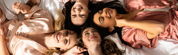 Vista superior de mulheres multiétnicas sorrindo e descansando na cama na festa de despedida de solteira, tiro panorâmico — Fotografia de Stock