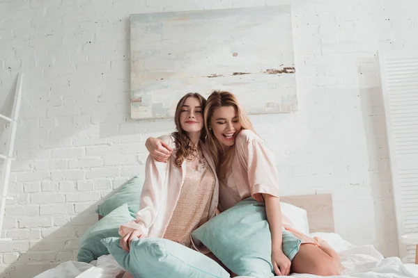 Attractive women hugging and smiling with pillows on bed at bachelorette party — Stock Photo