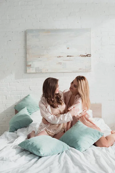 Mujeres atractivas abrazándose, sonriendo y mirándose con almohadas en la cama en la despedida de soltera - foto de stock