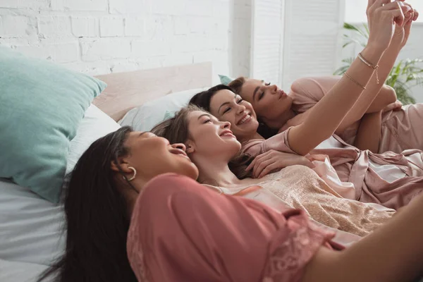 Selektiver Fokus aufgeregter und glücklicher multikultureller Frauen, die ein Selfie machen, während sie sich auf der Junggesellenparty im Bett ausruhen — Stockfoto