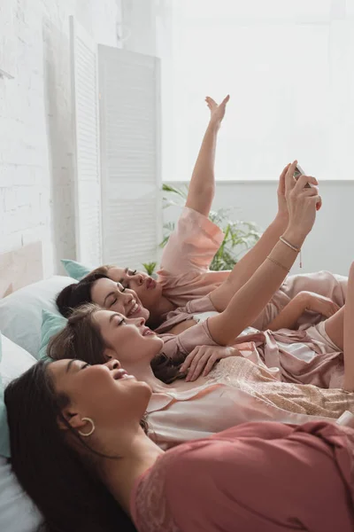 Selektiver Fokus multikultureller Frauen, die Selfie machen und auf der Junggesellenparty im Bett liegen — Stockfoto