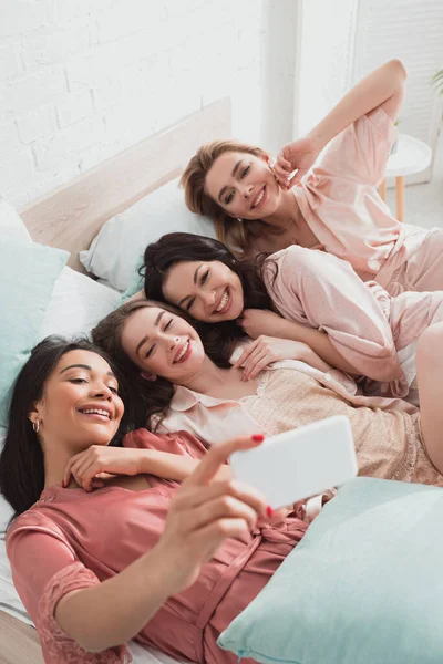 Focus selettivo della ragazza afro-americana che si fa selfie con gli amici mentre si sdraia sul letto all'addio al nubilato — Foto stock