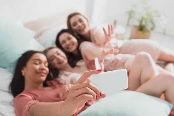 Selektiver Fokus eines afrikanisch-amerikanischen Mädchens, das Selfie mit Freunden macht, während es sich auf der Junggesellenabschiedsparty im Bett ausruht — Stockfoto