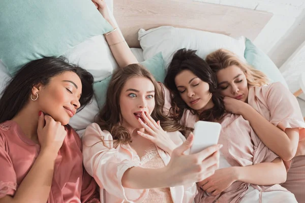 Blonde girl taking selfie with sleeping multiethnic friends on bed at bachelorette party — Stock Photo