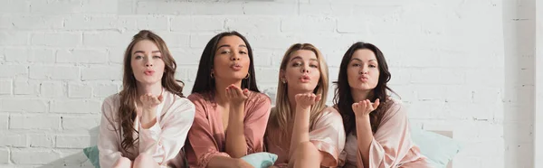 African american and caucasian women looking at camera and blowing kisses on bed at bachelorette party, panoramic shot — Stock Photo