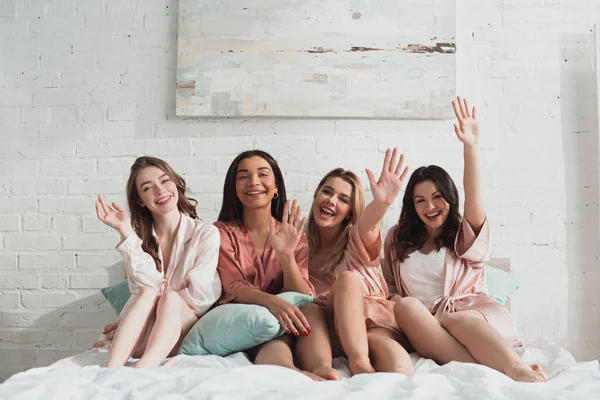 Multicultural women looking at camera, smiling and showing hello on bed at bachelorette party — Stock Photo