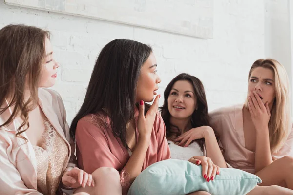 Multicultural women talking and looking at each other with pillow on bed at bachelorette party — Stock Photo