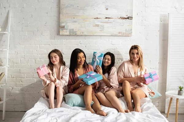 Multicultural women smiling and holding gift boxes on bed at bachelorette party — Stock Photo