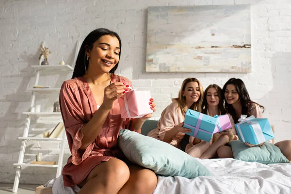 Enfoque selectivo de la chica afroamericana abriendo regalo con amigos sosteniendo cajas de regalo en la cama en la despedida de soltera - foto de stock