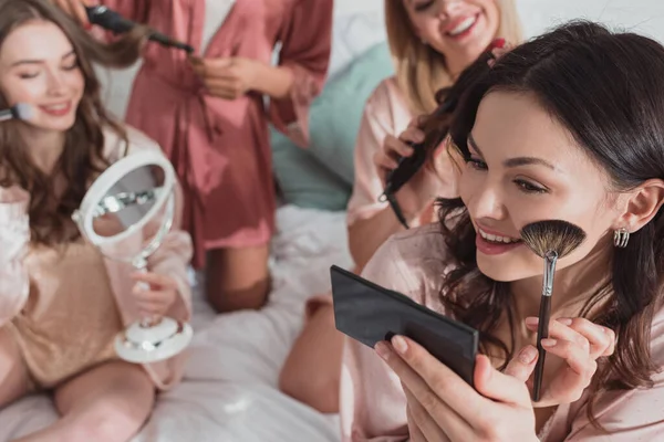 Vista cortada de amigos multiculturais colocando maquiagem e fazendo penteados com ferros de ondulação na cama — Fotografia de Stock