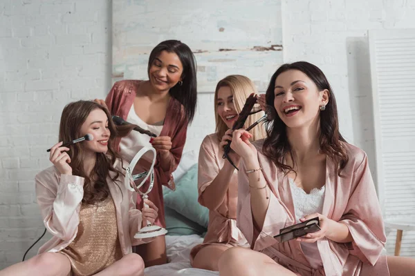 Multicultural friends putting makeup and doing hairstyles with curling irons at bachelorette party on bed — Stock Photo