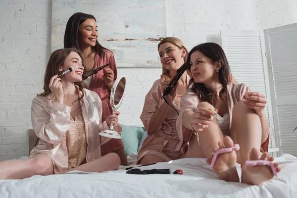 Селективный фокус мультикультурных женщин, накладывающих макияж и делающих прически на девичнике на кровати — стоковое фото