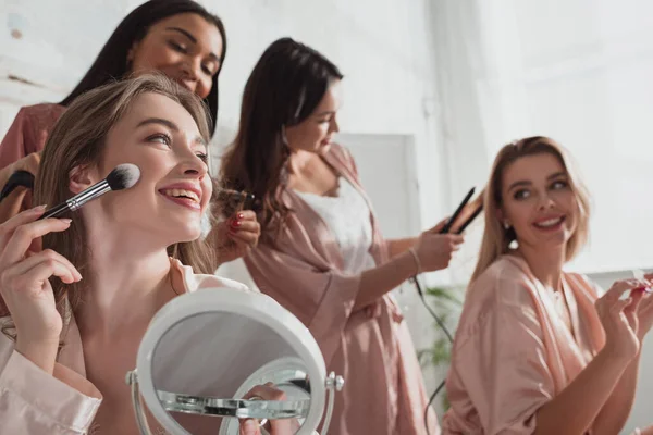 Foco seletivo de mulheres multiétnicas colocando maquiagem com escova cosmética e fazendo penteados no quarto na festa de despedida de solteira — Fotografia de Stock