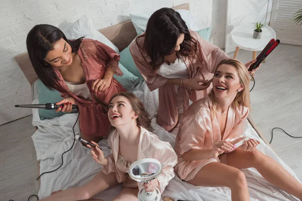Високий кут зору багатоетнічних жінок щасливі разом, використовуючи косметику і роблячи зачіски на ліжку — стокове фото