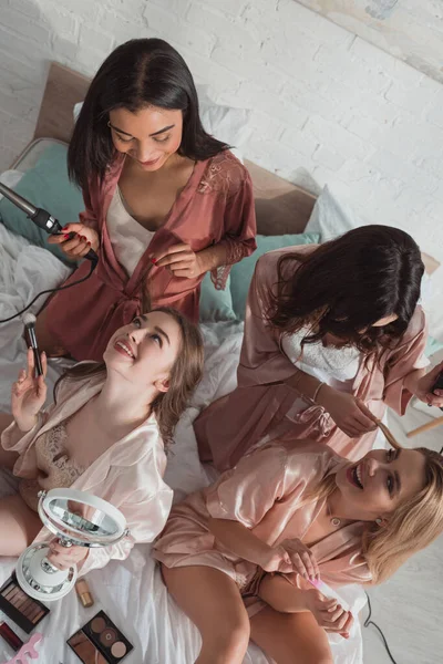 Vista ad alto angolo di donne multietniche che usano cosmetici e fanno acconciature all'addio al nubilato sul letto — Foto stock