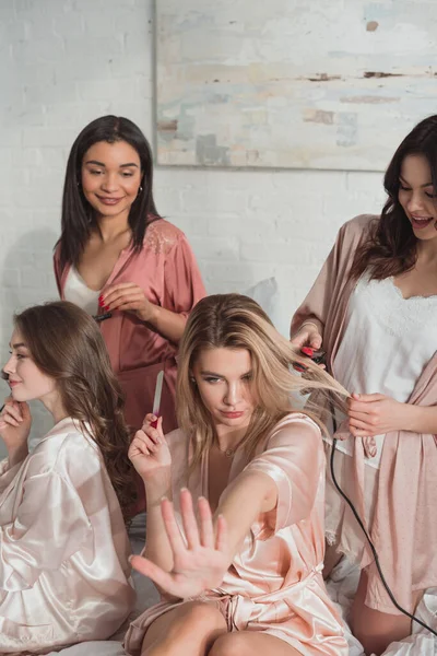 Blonde girl with nail file looking at manicure with multiethnic friends on bed at bachelorette party — Stock Photo