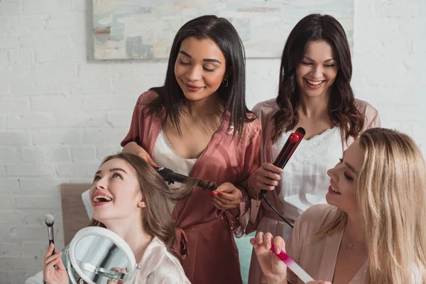 Multicultural friends doing hairstyles with curling irons and smiling at bachelorette party — Stock Photo