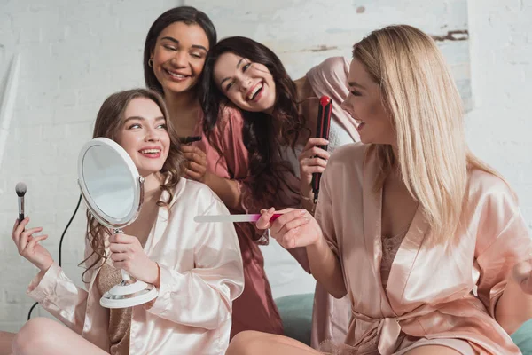 Mulheres multiculturais felizes com escova cosmética, arquivo de unhas e ferro de ondulação olhando um para o outro e sorrindo — Fotografia de Stock