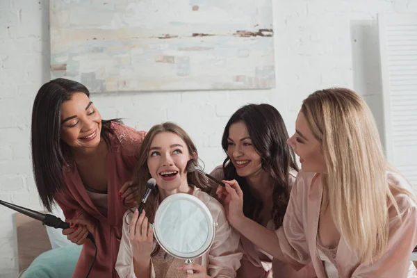 Mujeres multiculturales poniendo maquillaje en novia con cepillos cosméticos y sosteniendo espejo y rizador en despedida de soltera - foto de stock