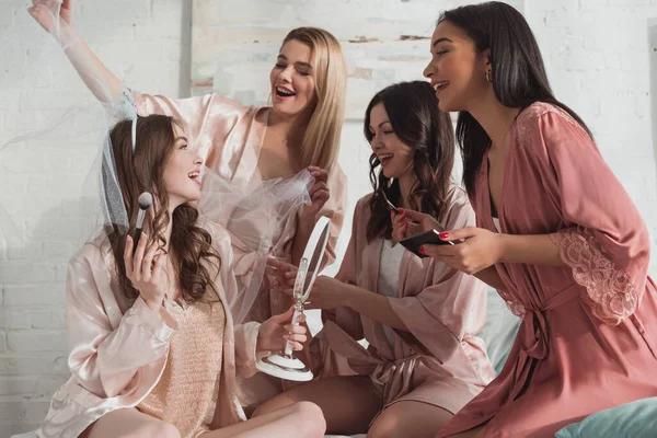 Feliz multiétnicas mujeres poniendo maquillaje en la novia en la habitación en la despedida de soltera - foto de stock