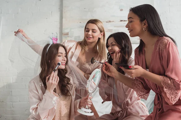 Las mujeres multiétnicas poniendo maquillaje en la novia juntos en la habitación en la despedida de soltera - foto de stock