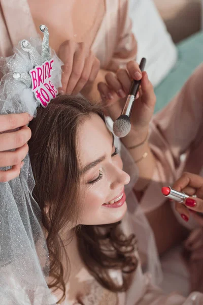 Vista recortada de mujeres multiculturales que ponen maquillaje con cepillo cosmético y lápiz labial en la novia en la despedida de soltera - foto de stock