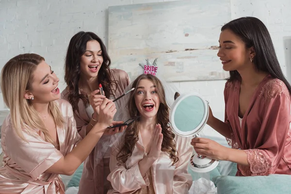 Mulheres multiétnicas colocando maquiagem com pincéis cosméticos em noiva animada e feliz com véu de noiva na festa de despedida de solteira — Fotografia de Stock