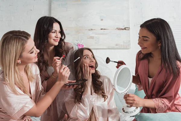 Multiethnic women putting makeup with cosmetic brushes on excited and happy bride at bachelorette party — Stock Photo