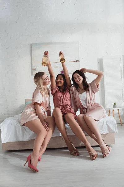 Happy multiethnic friends with bottles of champagne at bachelorette party on bed — Stock Photo